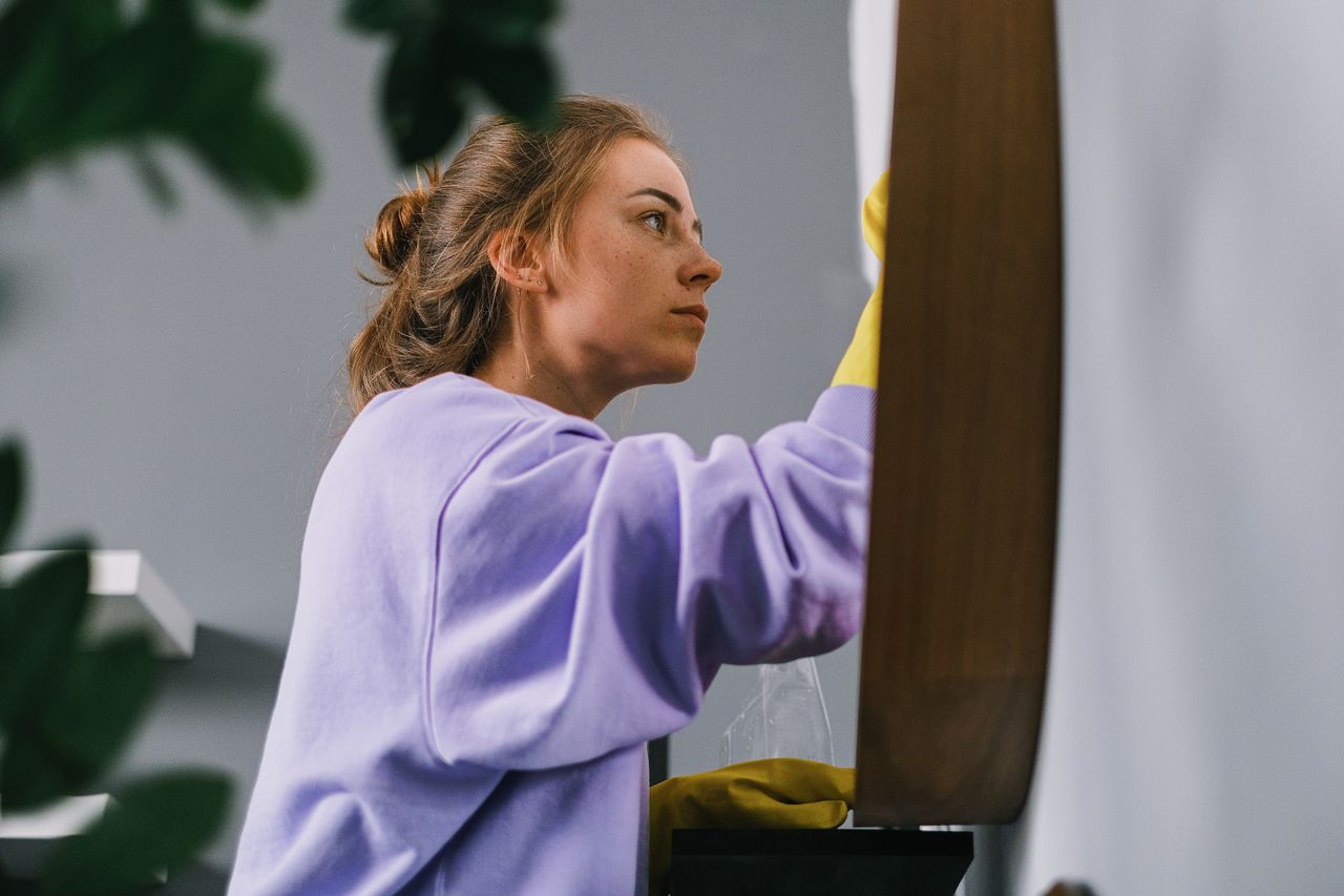 about-us-02 Side view of young focused female in latex gloves standing and cleaning mirror in light room in daytime